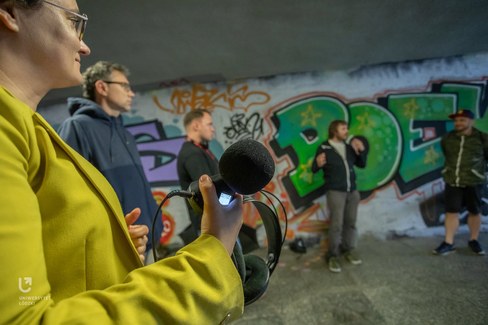 The photo shows a group of people standing in an underpass. They are holding audio recording devices.