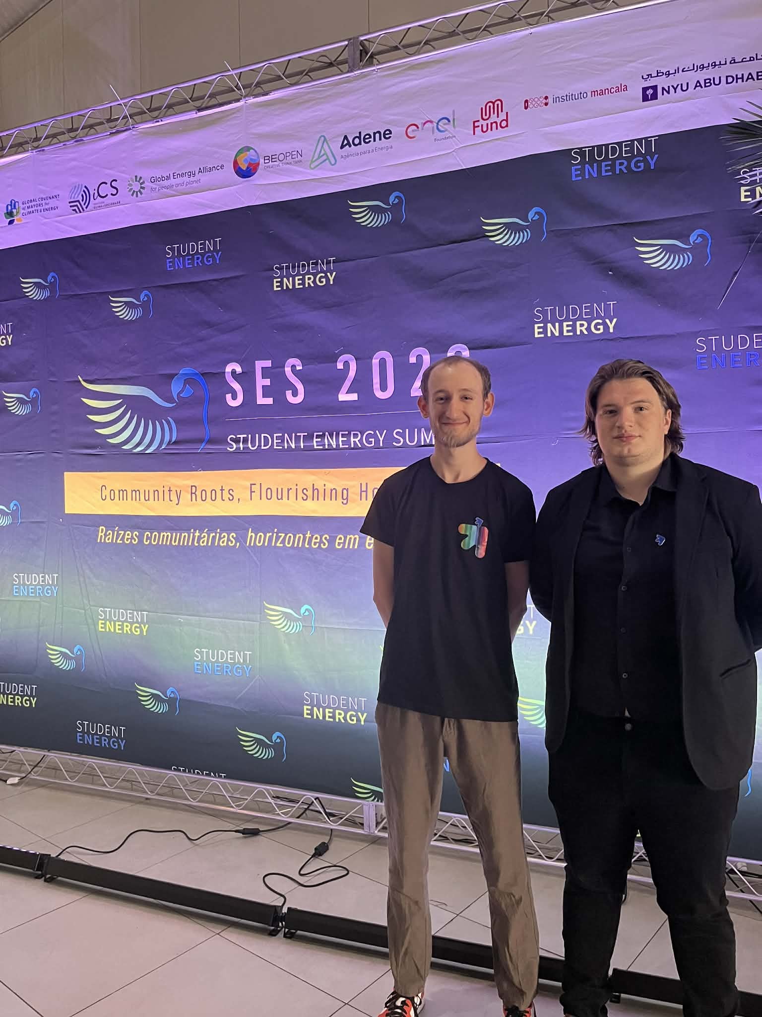 Dwie osoby stoją przed dużą planszą wydarzenia SES 2023 Student Energy Summit. Na banerze widnieje hasło ‘Community Roots, Flourishing Horizons’ oraz jego portugalska wersja, a także liczne logotypy partnerów i sponsorów. Scena znajduje się w jasnym, wewnętrznym pomieszczeniu z oświetleniem ustawionym przy podłodze