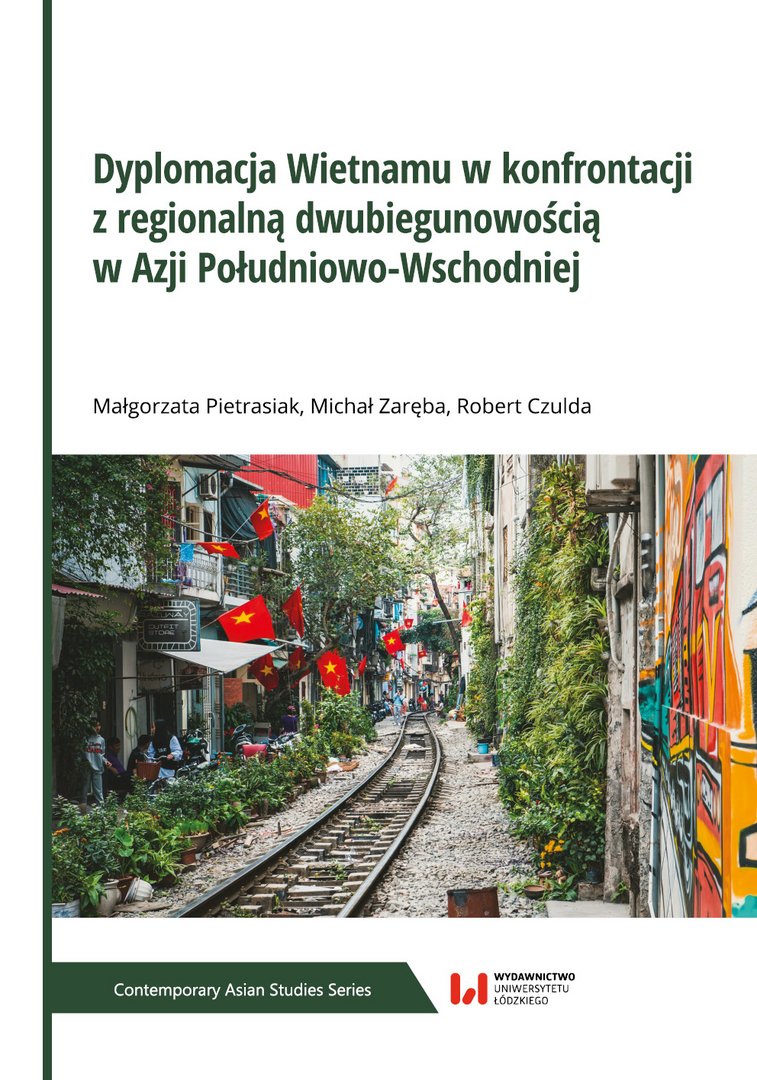 Okładka książki „Dyplomacja Wietnamu w konfrontacji z regionalną dwubiegunowością w Azji Południowo-Wschodniej”. Na okładce widoczna jest wąska kolejowa uliczka w gęsto zabudowanej części miasta w Wietnamie, z budynkami ozdobionymi flagami Wietnamu, roślinnością i muralami.