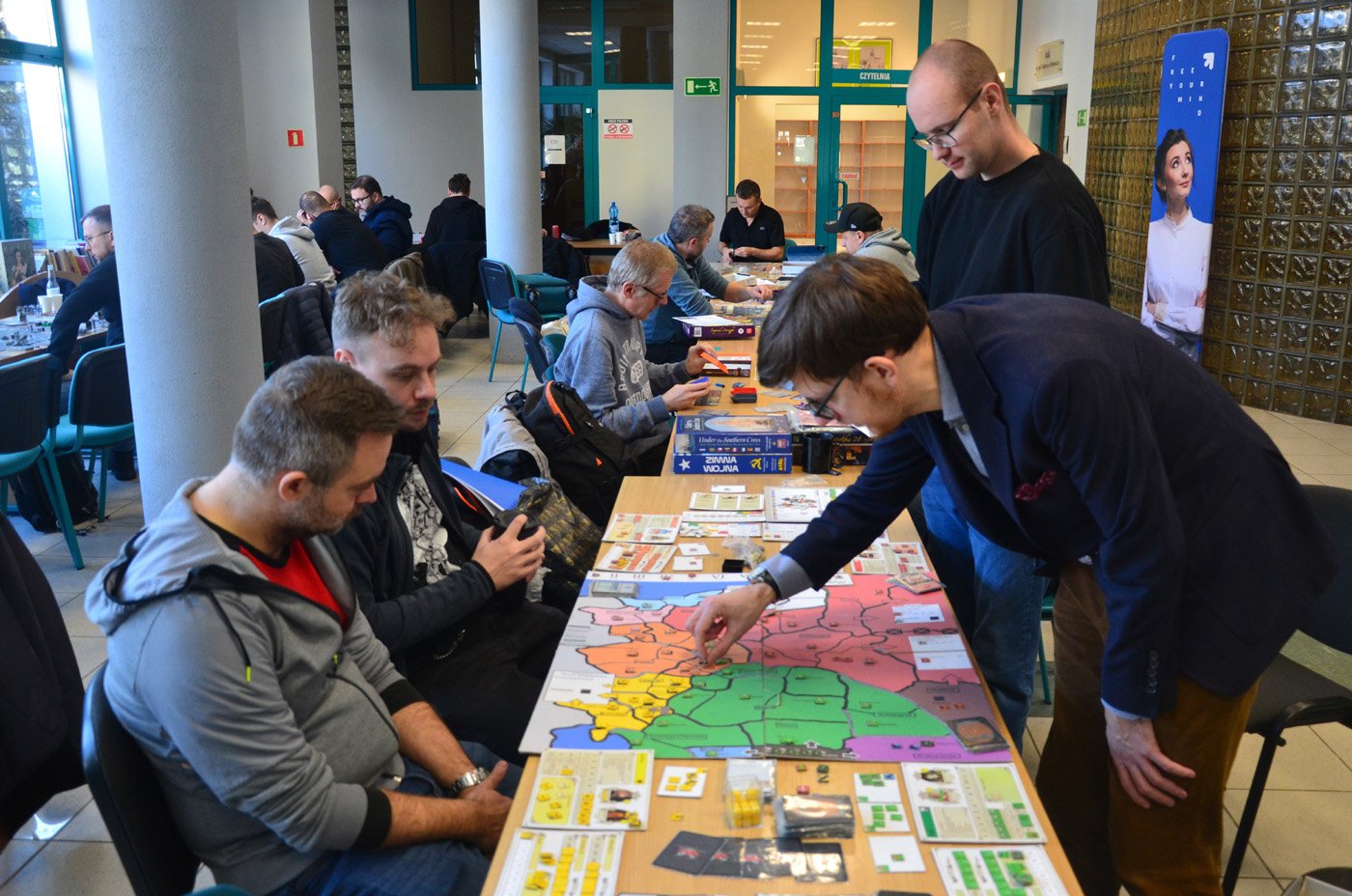 Grupa osób grających w gry planszowe w przestrzeni publicznej, prawdopodobnie w bibliotece lub centrum kultury. Na pierwszym planie cztery osoby skupione nad kolorową planszą z mapą, kartami i żetonami. Jedna z nich wykonuje ruch, umieszczając element na planszy. W tle widoczne są krzesła, stoły oraz plakat z napisem „Let your mind fly”. Atmosfera wydarzenia jest społeczna i rekreacyjna.