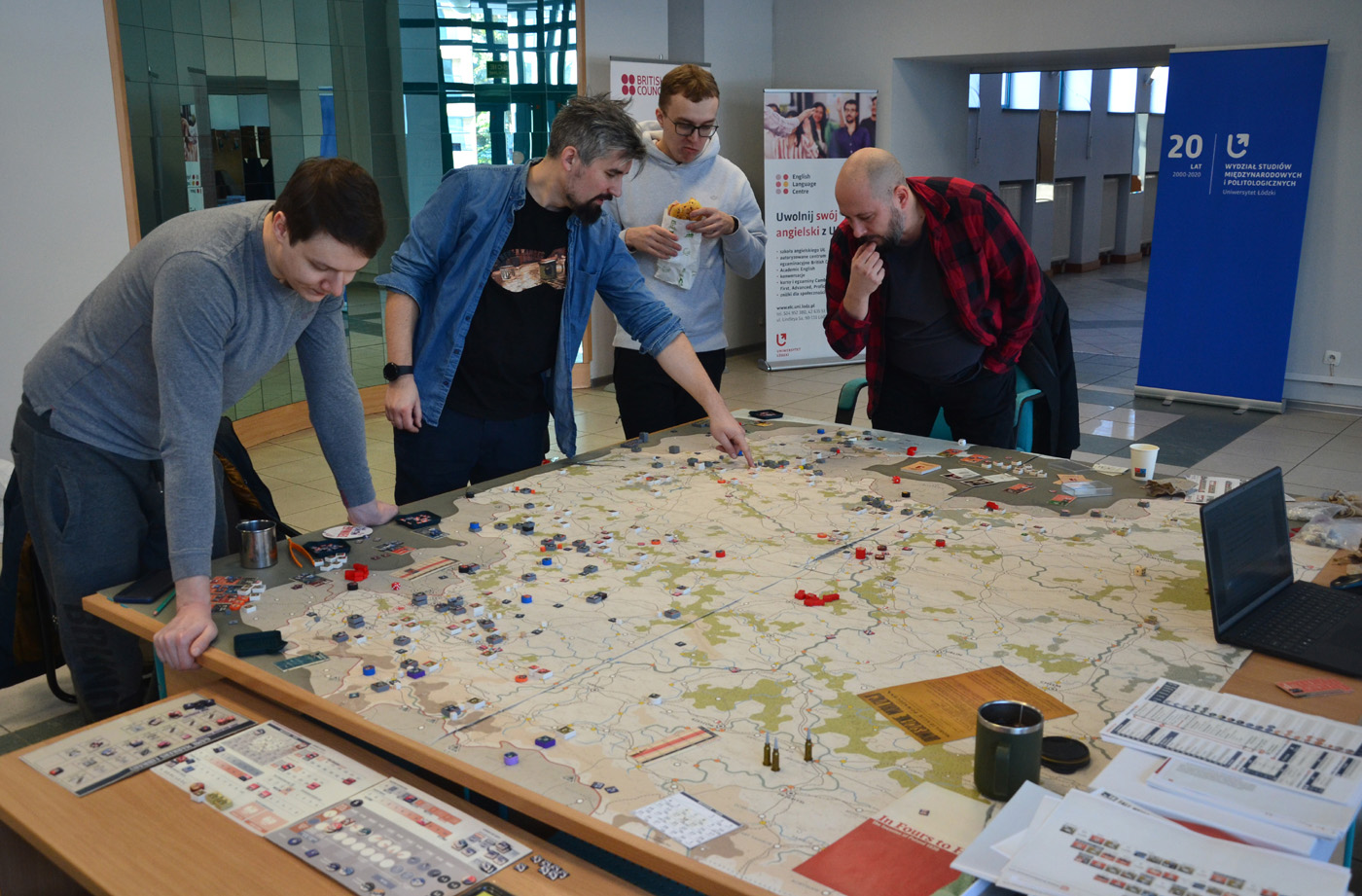 Pięć osób grających w strategiczną grę planszową przy stole w przestrzeni eventowej. Na stole rozłożona jest szczegółowa plansza, figurki, karty, kostki i pojemniki z żetonami. Jedna osoba wskazuje na planszę, pozostałe uważnie obserwują lub dyskutują. W tle widoczne są inne stoły z uczestnikami oraz pudełko gry „Successors”, sugerujące tematykę historyczną. Atmosfera skupienia i współpracy.