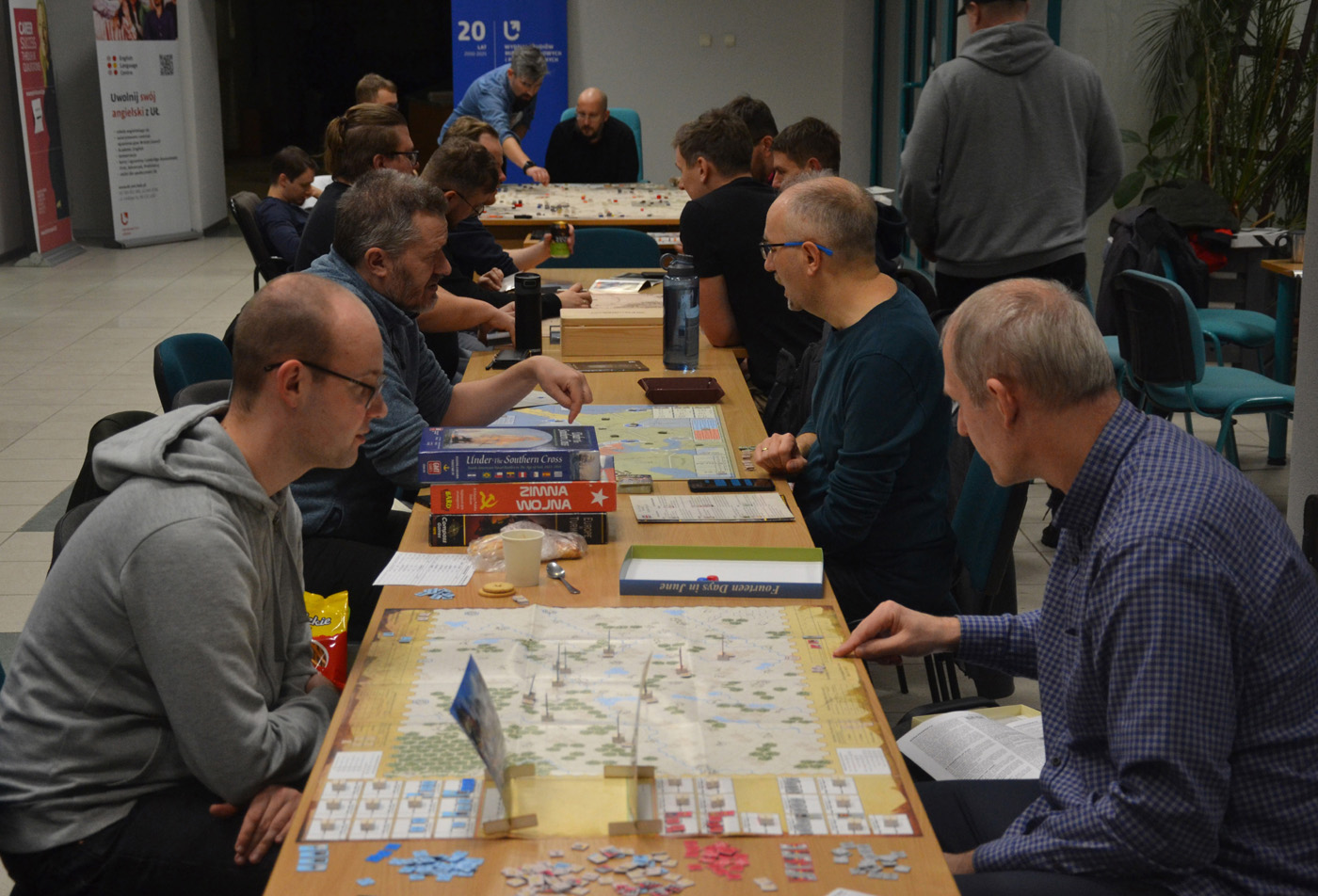Uczestnicy wydarzenia planszówkowego grają przy kilku stołach w strategiczne gry wojenne. Na stołach rozłożone są plansze z mapami, żetony, karty i pudełka gier, m.in. „Lions of Judah”, „Anzio”, „Under the Southern Cross” i „Fortress Rabaul”. Gracze są skupieni, a w tle widoczne są banery promocyjne i plakaty, co sugeruje zorganizowany charakter spotkania.