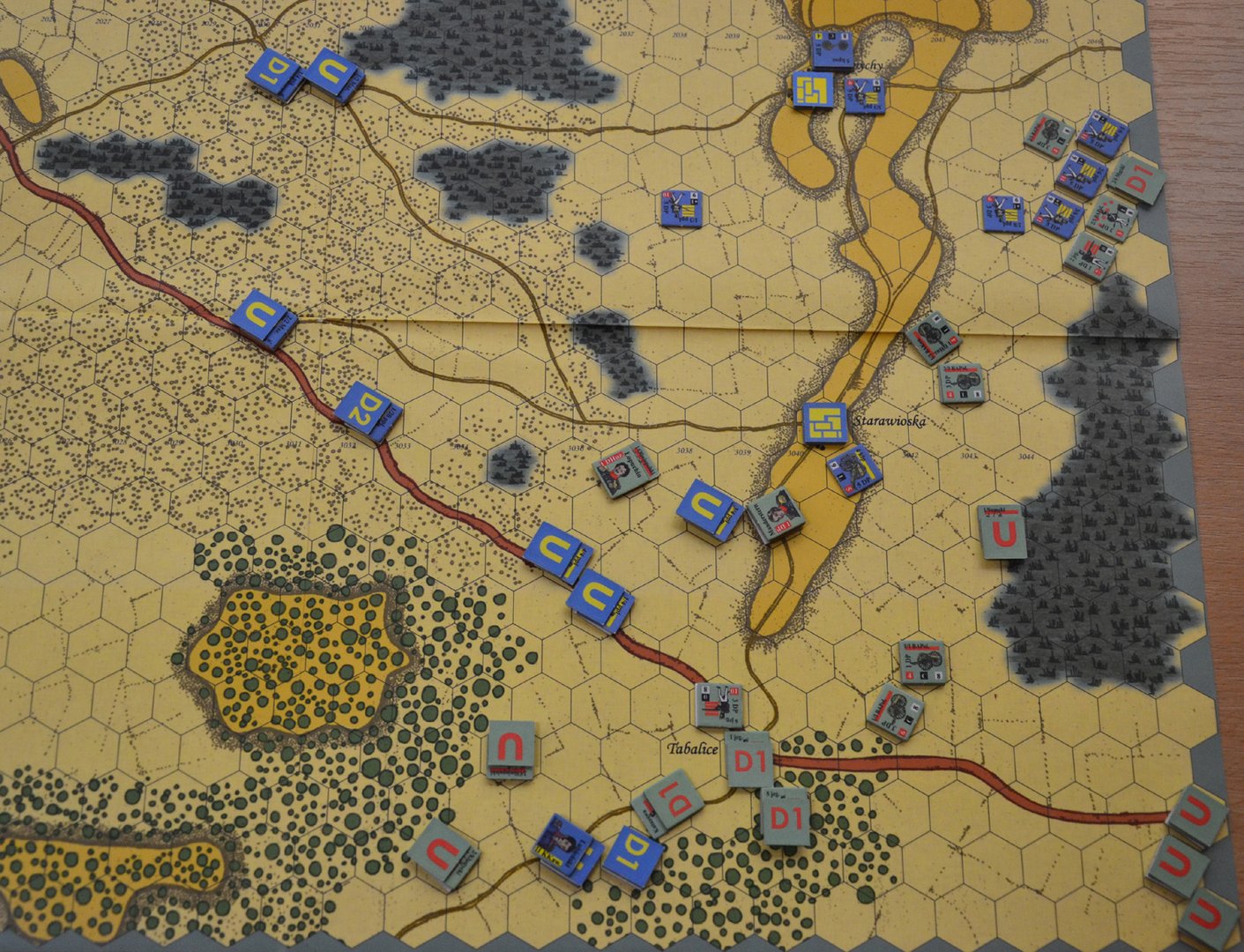Mapa strategicznej gry planszowej z siatką heksagonalną, przedstawiająca teren z lasami, drogami i wzniesieniami. Na mapie widoczne są oznaczone miejscowości, m.in. „Starawioska” i „Tabalice”. Rozmieszczone są kolorowe żetony jednostek z literami i symbolami, reprezentujące różne frakcje wojskowe — m.in. niebieskie i żółte z oznaczeniami „C” i „P” oraz szare i czerwone z „U” i „D1”. Układ sugeruje rozgrywkę symulującą działania wojenne.