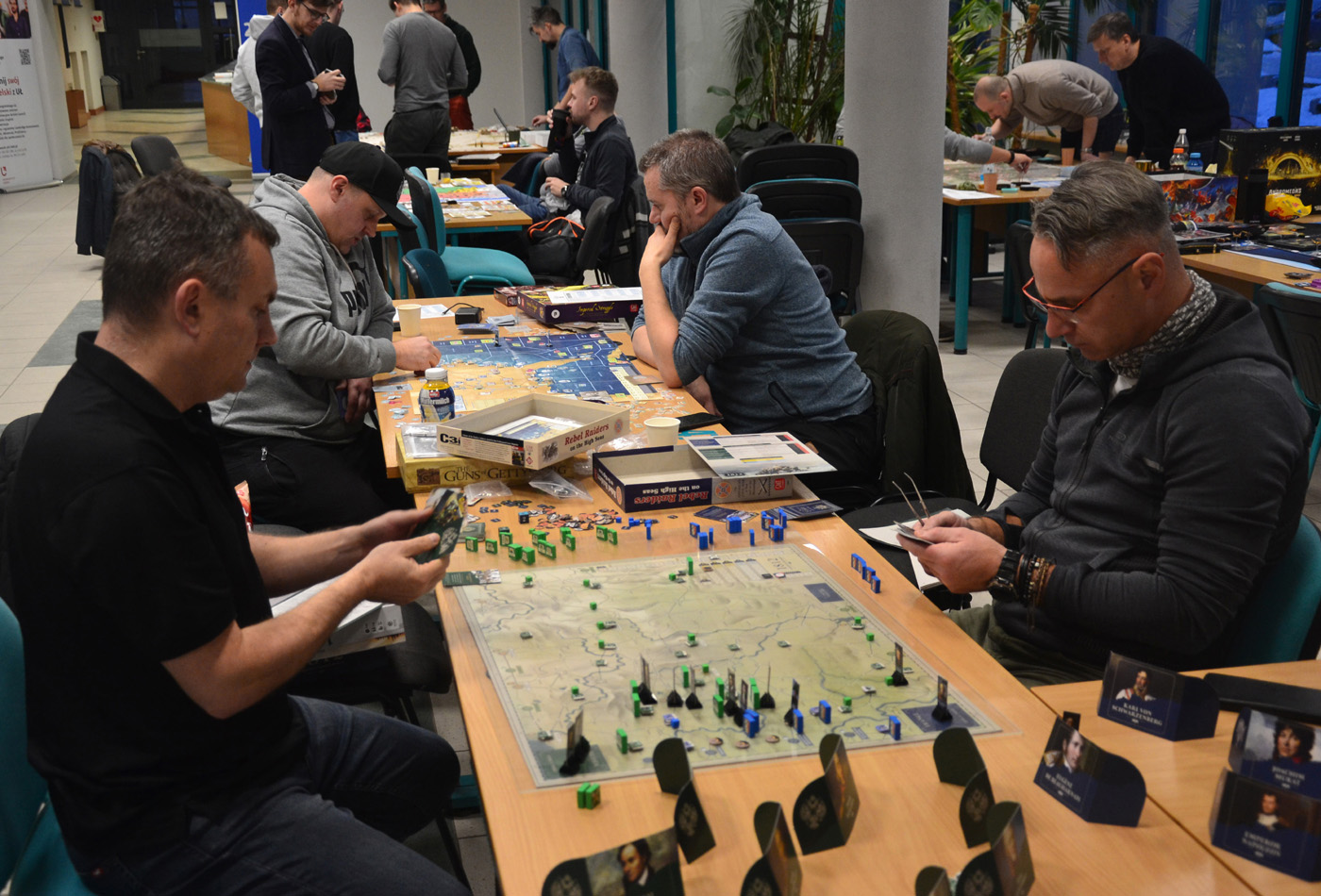 Uczestnicy wydarzenia planszówkowego grają przy kilku stołach w zaawansowane gry strategiczne. Na pierwszym planie widoczna jest plansza z mapą, zielonymi i niebieskimi znacznikami oraz kartami postaci z portretami. W tle znajdują się inne stoły z graczami oraz pudełka gier, m.in. „Guns of Gettysburg”, „Rebel Raiders on the High Seas” i „Liberty or Death”. Atmosfera skupienia i zaangażowania.