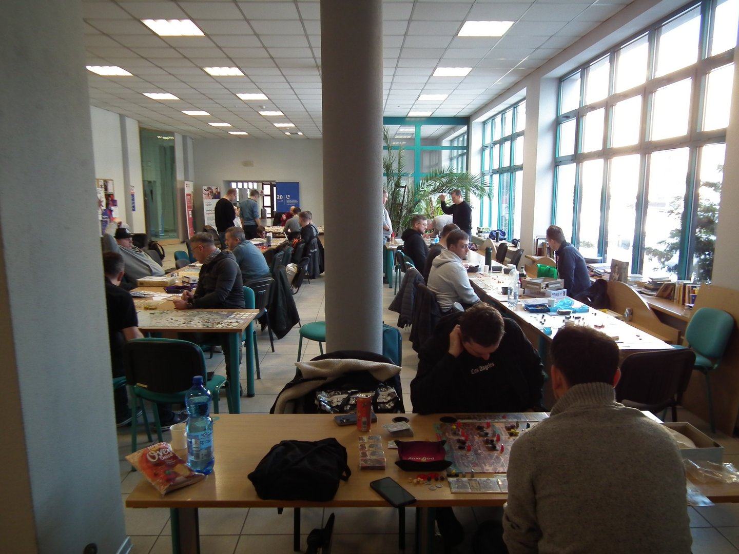 Sala pełna uczestników grających w gry planszowe. Przy kilku stołach siedzą grupy osób skupionych na rozgrywce strategicznej. Na stołach widoczne są plansze, karty, pionki i pudełka gier. Atmosfera jest towarzyska i swobodna — niektóre stoły mają przekąski i napoje. W tle widać plakaty i banery, co sugeruje zorganizowane wydarzenie, np. turniej lub konwent.