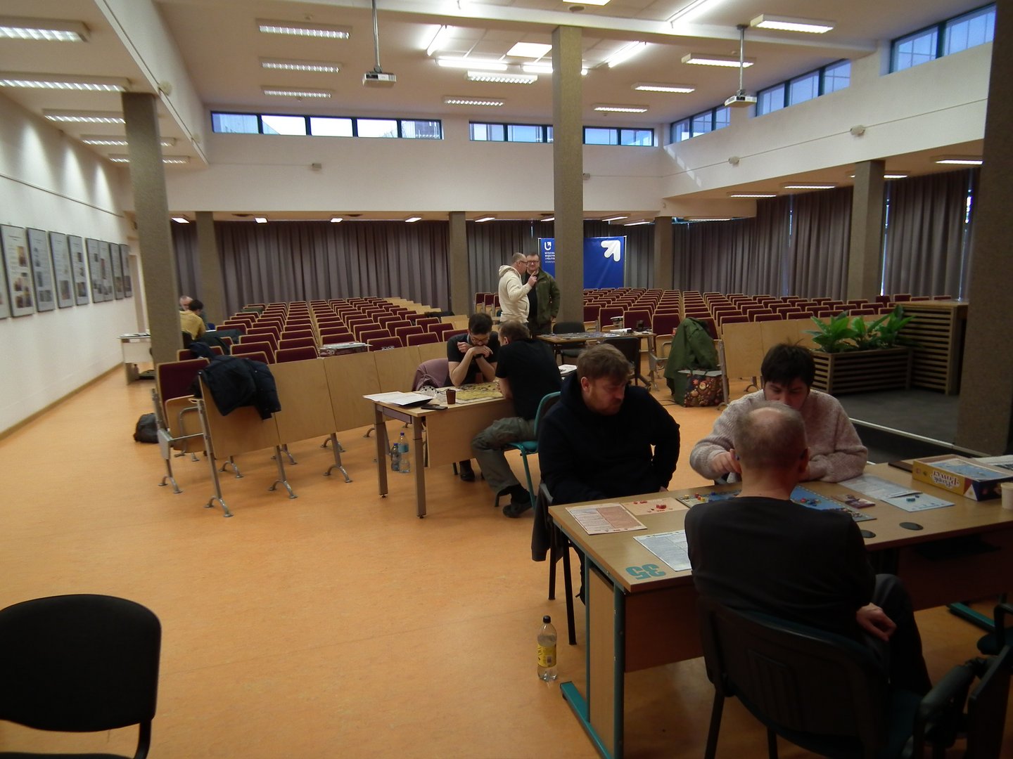 Widok sali z czerwonymi krzesłami ustawionymi w rzędach w stylu konferencyjnym, skierowanymi w stronę sceny z niebieskim banerem. Na pierwszym planie dwa stoły, przy których uczestnicy grają w gry planszowe — widoczne są karty, pionki i pudełko gry „Dominion”. Atmosfera jest swobodna, a przestrzeń sugeruje wydarzenie o charakterze edukacyjnym lub rekreacyjnym.