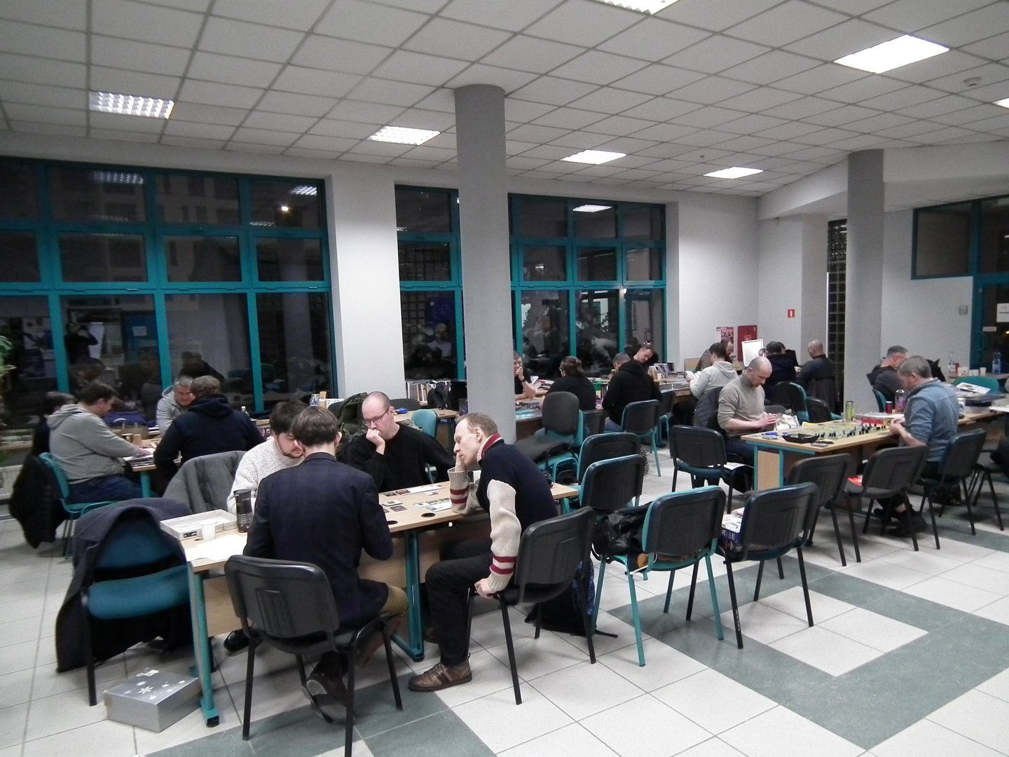 Sala z dużymi oknami i jasnym oświetleniem, wypełniona uczestnikami grającymi w gry planszowe. Przy kilku stołach siedzą grupy osób skupionych na rozgrywkach strategicznych lub modelarskich. Na stołach widoczne są plansze, figurki, pudełka gier i akcesoria. Atmosfera jest społeczna i rekreacyjna, sugerująca wydarzenie hobbystyczne lub spotkanie klubu gier.