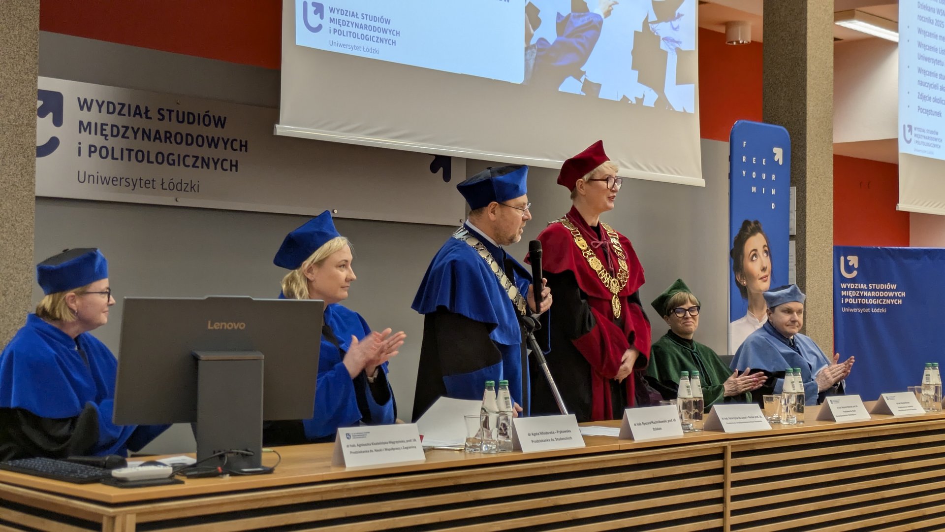 Uroczystość akademicka na Uniwersytecie Łódzkim. Za długim stołem siedzą osoby w togach, w tym dziekan w czerwonej szacie z łańcuchem. W tle widoczne są banery wydziału oraz ekran z nazwą jednostki.