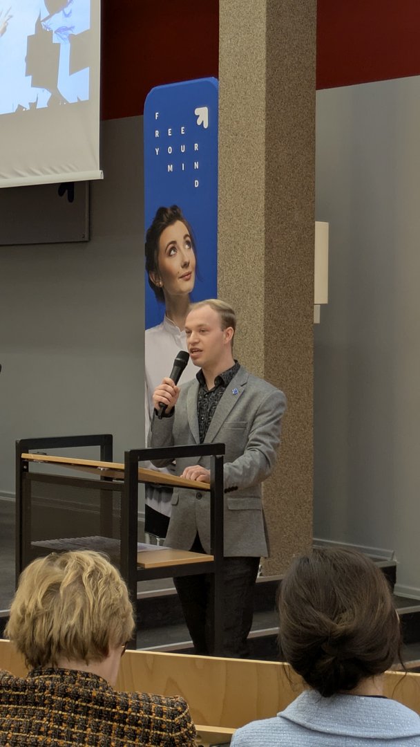 Mężczyzna w szarej marynarce przemawia przy mównicy podczas wydarzenia edukacyjnego. Za nim stoi pionowy baner z wizerunkiem kobiety i napisem ‘FREE YOUR MIND’. W sali siedzą słuchacze zwróceni w jego stronę.