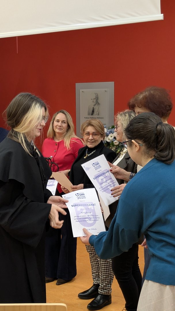 Osoba w czarnej todze wręcza certyfikaty kilku elegancko ubranym uczestnikom stojącym w rzędzie. W tle widoczna jest czerwona ściana z tablicą pamiątkową oraz osoby trzymające dokumenty z napisem ‘SUPERDZIEKANAT'