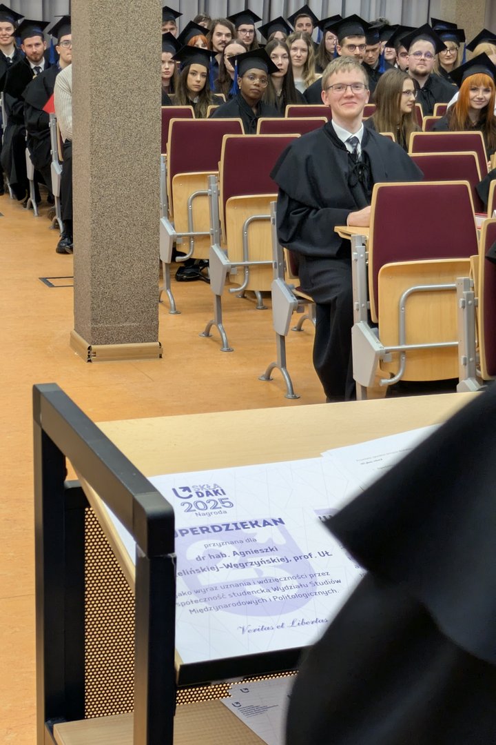 Studenci w czarnych togach siedzą w auli podczas uroczystości, a jedna osoba na pierwszym planie uśmiecha się do kamery. Na pulpicie przed nią leży widoczny fragment dyplomu z napisem ‘SKŁADAKI 2025'
