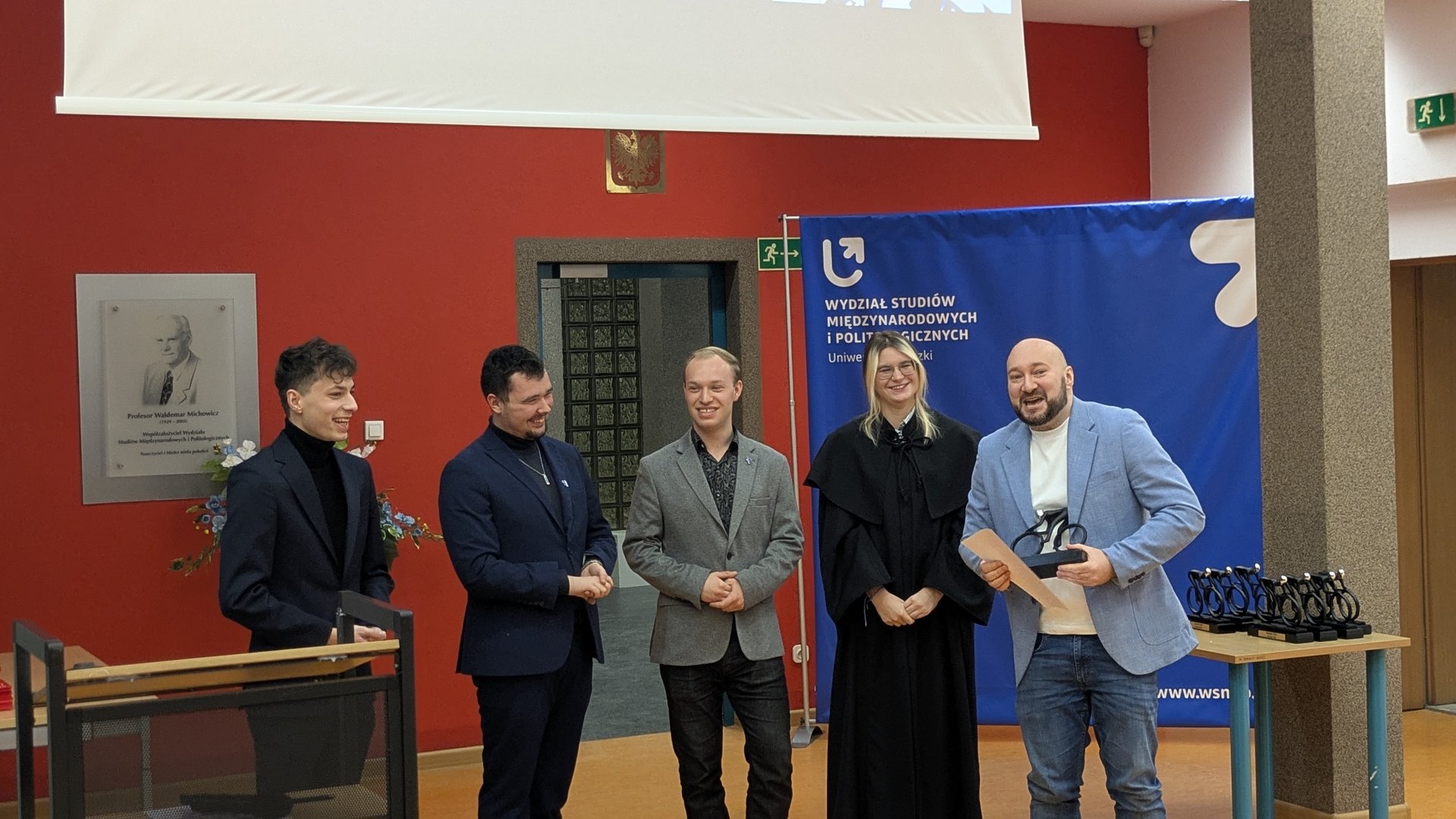 Pięć osób stoi w sali podczas uroczystości akademickiej. Jedna z nich ma na sobie togę, a mężczyzna po prawej trzyma puchar i dyplom. W tle widoczny jest niebieski baner wydziału Uniwersytetu Łódzkiego, czerwona ściana z tablicą pamiątkową oraz stół z nagrodami.