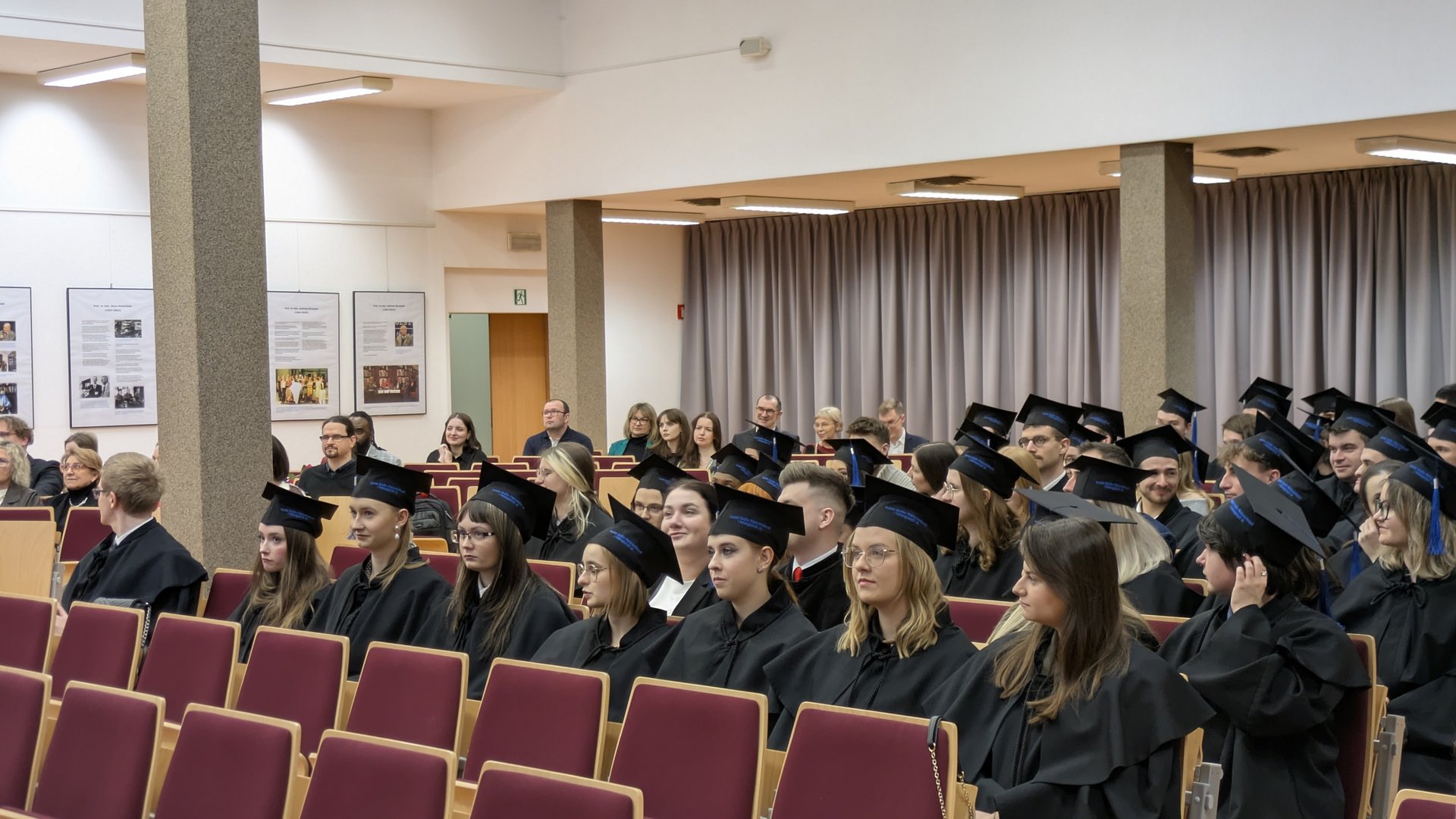 Absolwenci w czarnych togach siedzą w czerwonych fotelach podczas uroczystości w auli. W tle widoczni są goście i rodziny obserwujące wydarzenie, a na ścianach wiszą plakaty. Sala jest jasno oświetlona.
