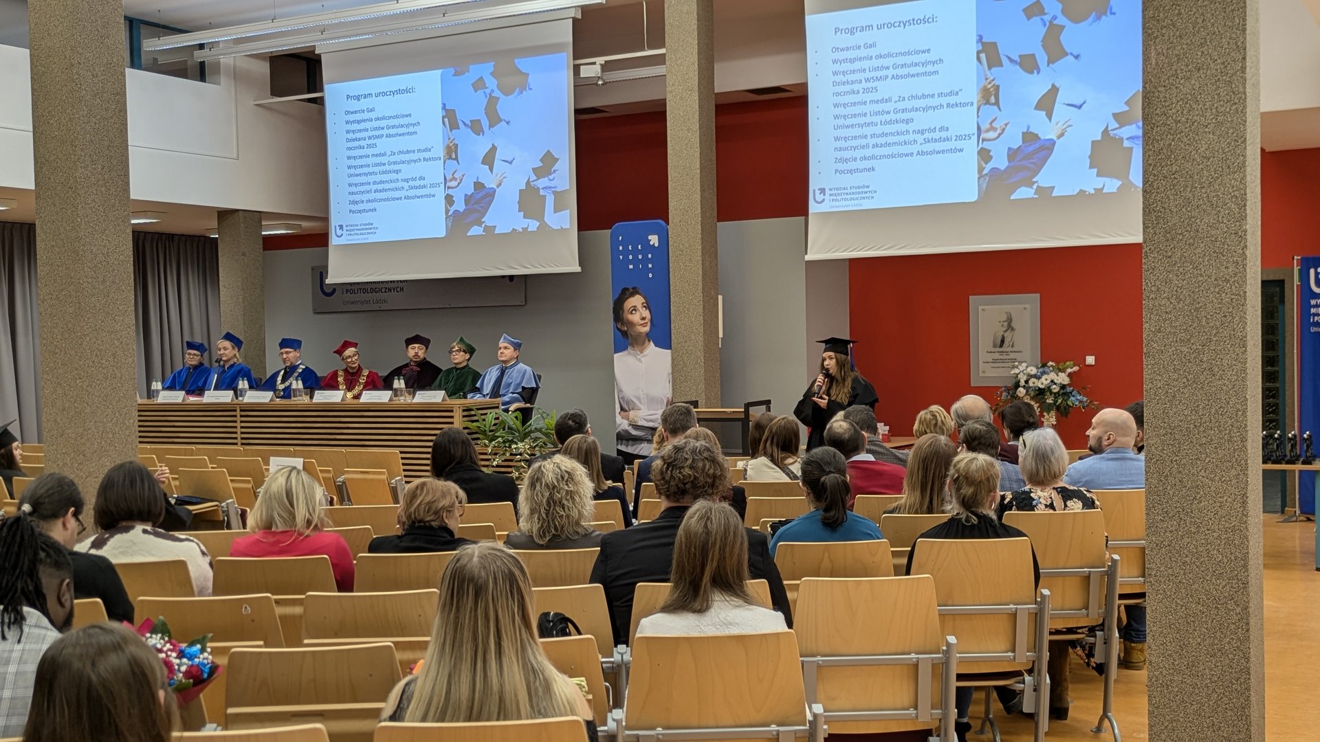 Uroczystość akademicka w auli Uniwersytetu Łódzkiego. Przy stole na scenie siedzą osoby w togach, a przed nimi publiczność. Studentka w todze przemawia z mównicy, a na dwóch ekranach wyświetlany jest program gali