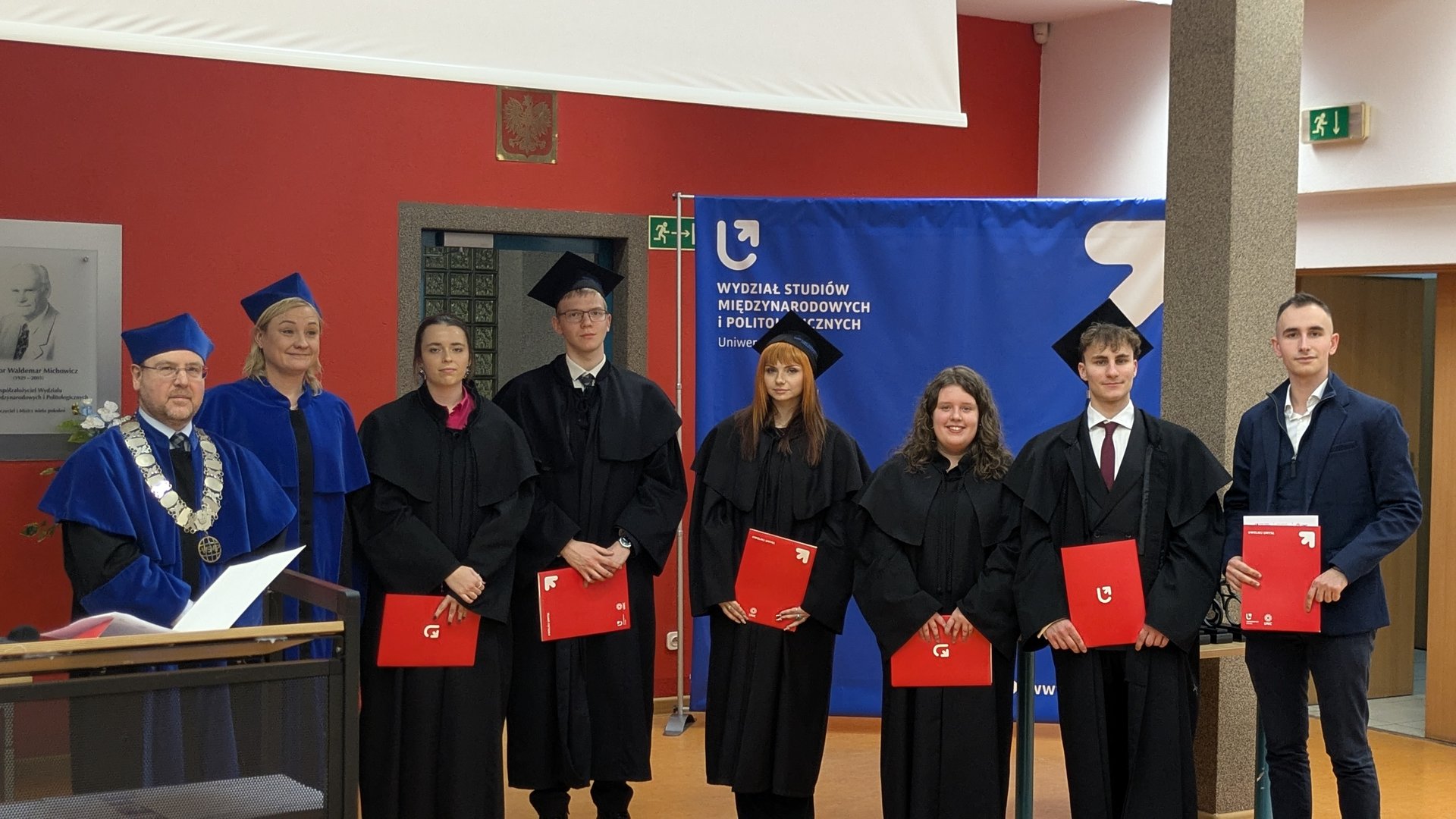 Osiem osób stoi w rzędzie podczas uroczystości na Uniwersytecie Łódzkim. Pięciu absolwentów w czarnych togach trzyma czerwone teczki, obok nich stoją dwaj wykładowcy w niebieskich szatach oraz jedna osoba w garniturze. W tle widoczny jest niebieski baner wydziału i tablica pamiątkowa