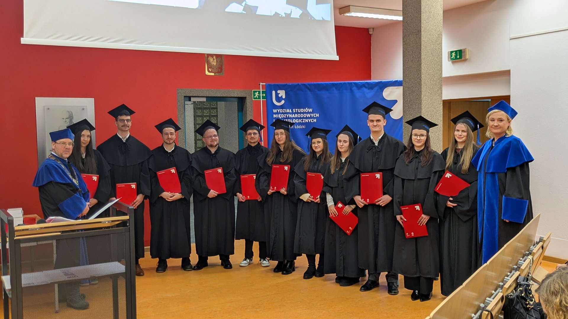 Grupa absolwentów w czarnych togach stoi w rzędzie, każdy trzyma czerwoną teczkę z dyplomem. Po obu stronach stoją wykładowcy w niebieskich szatach. W tle widoczny jest czerwony fragment ściany, niebieski baner wydziału Uniwersytetu Łódzkiego oraz ekran projekcyjny