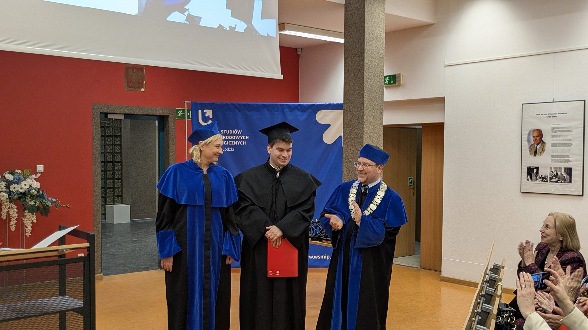 Absolwent w czarnej todze stoi między dwoma wykładowcami w niebieskich szatach; trzyma czerwoną teczkę z dyplomem. W tle widoczny jest niebieski baner wydziału Uniwersytetu Łódzkiego, ekran projekcyjny oraz tablica pamiątkowa. Kilka osób siedzi i bije brawo.
