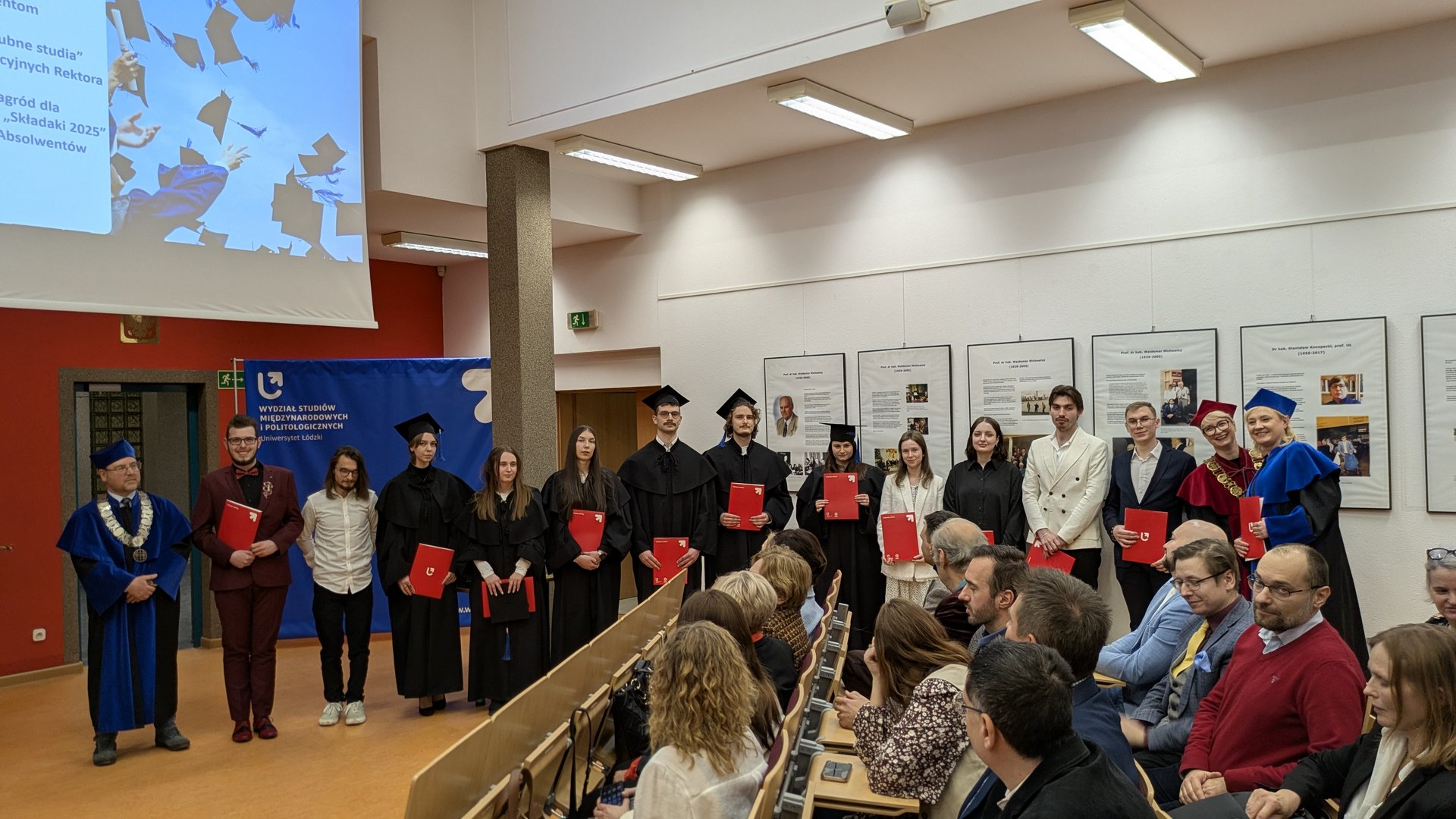 Grupa absolwentów w togach stoi w rzędzie, trzymając czerwone teczki z dyplomami. Obok nich stoją wykładowcy w uroczystych szatach, w tym jeden z łańcuchem dziekańskim. W tle widoczny jest ekran z grafiką zrzucanych biretów oraz niebieski baner wydziału Uniwersytetu Łódzkiego. Publiczność siedzi naprzeciw sceny