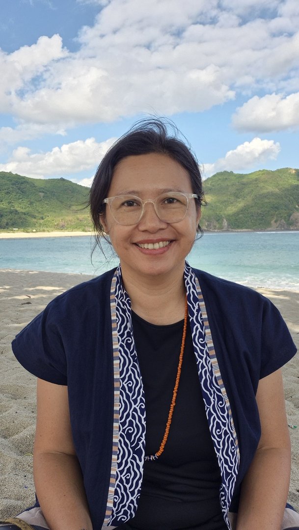 Dian Purnomo sitting on a sandy beach with a view of turquoise sea and green hills in the background, under a partly cloudy sky. Dressed in a dark shirt, a blue-and-white patterned scarf, and an orange beaded necklace, she wears glasses and smiles at the camera. The scenery suggests a tropical or coastal vacation spot. Dian Purnomo sitting on a sandy beach with a view of turquoise sea and green hills in the background, under a partly cloudy sky. Dressed in a dark shirt, a blue-and-white patterned scarf, and an orange beaded necklace, she wears glasses and smiles at the camera. The scenery suggests a tropical or coastal vacation spot.