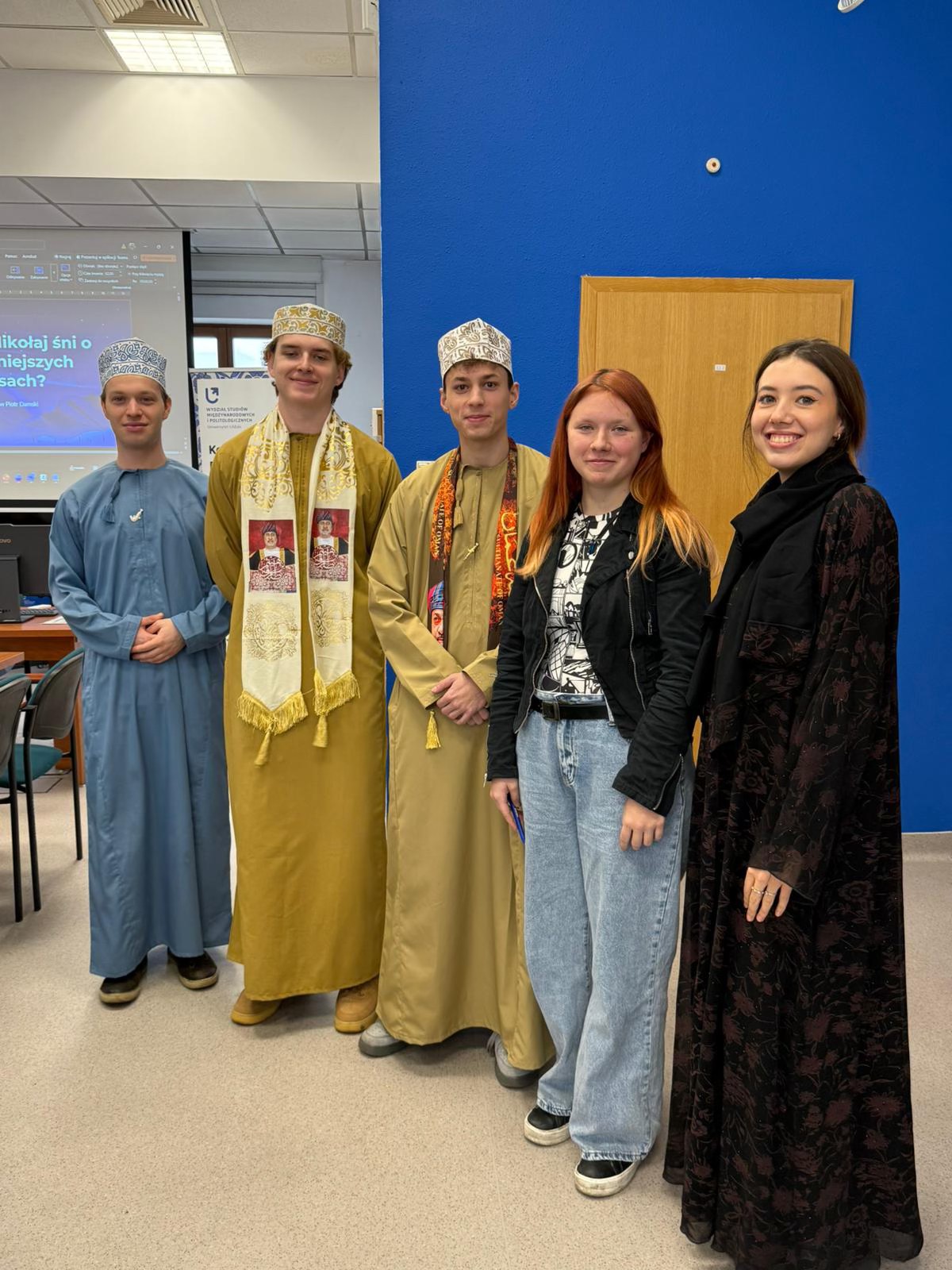 Pięć osób stoi w pomieszczeniu przed niebieską ścianą i drewnianymi drzwiami. Trzy osoby mają na sobie tradycyjne stroje bliskowschodnie, w tym długie szaty i wzorzyste nakrycia głowy. Dwie pozostałe są ubrane w nowoczesne, codzienne ubrania. W tle widoczny jest ekran z prezentacją w języku polskim. Pięć osób stoi w pomieszczeniu przed niebieską ścianą i drewnianymi drzwiami. Trzy osoby mają na sobie tradycyjne stroje bliskowschodnie, w tym długie szaty i wzorzyste nakrycia głowy. Dwie pozostałe są ubrane w nowoczesne, codzienne ubrania. W tle widoczny jest ekran z prezentacją w języku polskim.