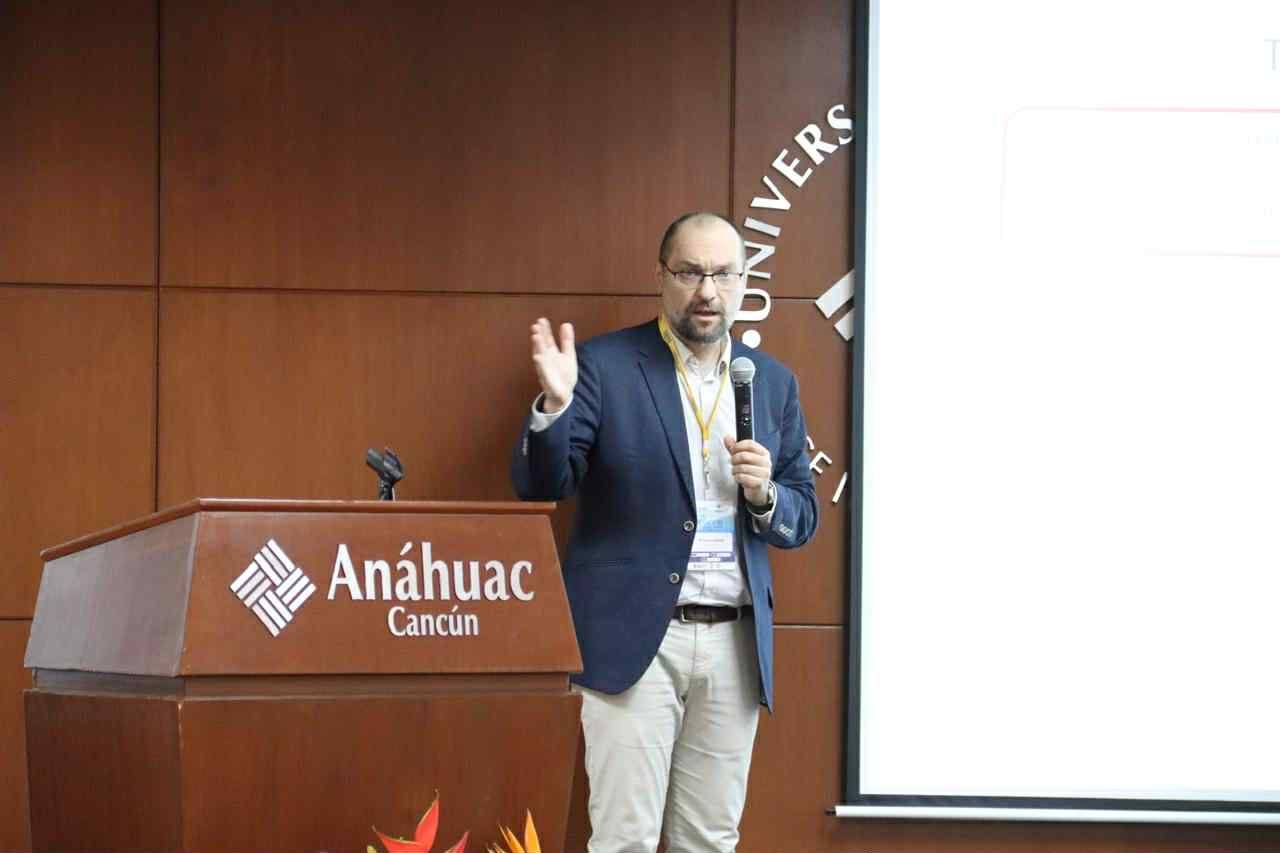 prof. Tomasz Kamiński prowadzi prezentację na Uniwersytecie Anáhuac Cancún. Stoi przed mównicą z nazwą uczelni, trzyma mikrofon w lewej ręce i gestykuluje prawą. Ubrany jest w okulary, ciemną marynarkę, jasną koszulę i beżowe spodnie. Za nim znajduje się ekran projekcyjny z częściowo niewyraźnym slajdem oraz ściana z logo i nazwą uczelni. Zdjęcie uchwyca moment akademickiego wystąpienia, podczas konferencji prof. Tomasz Kamiński prowadzi prezentację na Uniwersytecie Anáhuac Cancún. Stoi przed mównicą z nazwą uczelni, trzyma mikrofon w lewej ręce i gestykuluje prawą. Ubrany jest w okulary, ciemną marynarkę, jasną koszulę i beżowe spodnie. Za nim znajduje się ekran projekcyjny z częściowo niewyraźnym slajdem oraz ściana z logo i nazwą uczelni. Zdjęcie uchwyca moment akademickiego wystąpienia, podczas konferencji