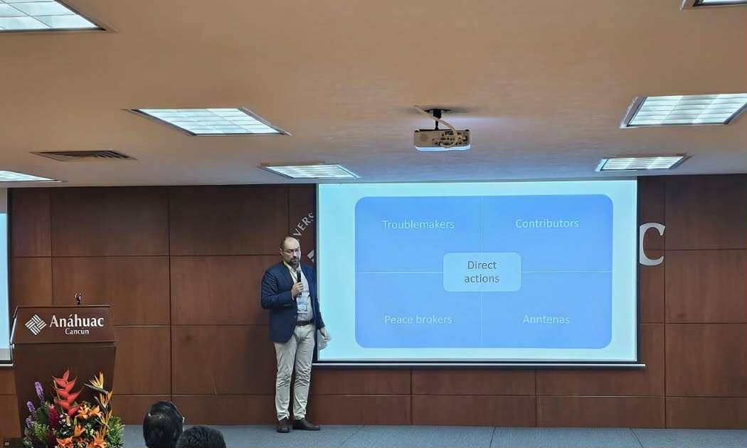 prof. Tomasz Kamiński prowadzi prezentację w sali konferencyjnej Uniwersytetu Anáhuac Cancún. Trzyma mikrofon i stoi obok ekranu projekcyjnego, na którym wyświetlony jest slajd z diagramem. Centralne pole diagramu nosi nazwę ‘Direct actions’, a wokół niego rozmieszczone są cztery sekcje: ‘Troublemakers’, ‘Contributors’, ‘Peace brokers’ i ‘Antennas’. W tle widoczna jest drewniana ściana z logo uczelni, a obok mównicy znajduje się kompozycja kwiatowa. Sufit wyposażony jest w oświetlenie punktowe i projektor. Zdjęcie przedstawia profesjonalne wystąpienie, prawdopodobnie dotyczące dynamiki zespołu lub ról organizacyjnych. prof. Tomasz Kamiński prowadzi prezentację w sali konferencyjnej Uniwersytetu Anáhuac Cancún. Trzyma mikrofon i stoi obok ekranu projekcyjnego, na którym wyświetlony jest slajd z diagramem. Centralne pole diagramu nosi nazwę ‘Direct actions’, a wokół niego rozmieszczone są cztery sekcje: ‘Troublemakers’, ‘Contributors’, ‘Peace brokers’ i ‘Antennas’. W tle widoczna jest drewniana ściana z logo uczelni, a obok mównicy znajduje się kompozycja kwiatowa. Sufit wyposażony jest w oświetlenie punktowe i projektor. Zdjęcie przedstawia profesjonalne wystąpienie, prawdopodobnie dotyczące dynamiki zespołu lub ról organizacyjnych.