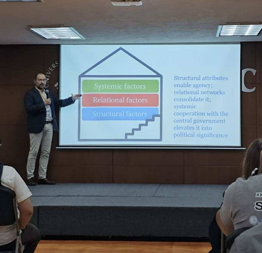 prof. Tomasz Kamiński prowadzi wykład lub prezentację, wskazując na slajd wyświetlony na ścianie za nim. Slajd przedstawia diagram domu z trzema kolorowymi sekcjami: zieloną ‘Systemic factors’, niebieską ‘Relational factors’ i czerwoną ‘Structural factors’. Przez środek diagramu biegną schody prowadzące ku górze. Po prawej stronie slajdu znajduje się tekst: ‘Structural attributes enable agency; relational networks consolidate it; systemic cooperation with the central government elevates it into political significance’. Zdjęcie ukazuje moment akademickiego wystąpienia, prawdopodobnie dotyczącego struktur społecznych lub politycznych. prof. Tomasz Kamiński prowadzi wykład lub prezentację, wskazując na slajd wyświetlony na ścianie za nim. Slajd przedstawia diagram domu z trzema kolorowymi sekcjami: zieloną ‘Systemic factors’, niebieską ‘Relational factors’ i czerwoną ‘Structural factors’. Przez środek diagramu biegną schody prowadzące ku górze. Po prawej stronie slajdu znajduje się tekst: ‘Structural attributes enable agency; relational networks consolidate it; systemic cooperation with the central government elevates it into political significance’. Zdjęcie ukazuje moment akademickiego wystąpienia, prawdopodobnie dotyczącego struktur społecznych lub politycznych.