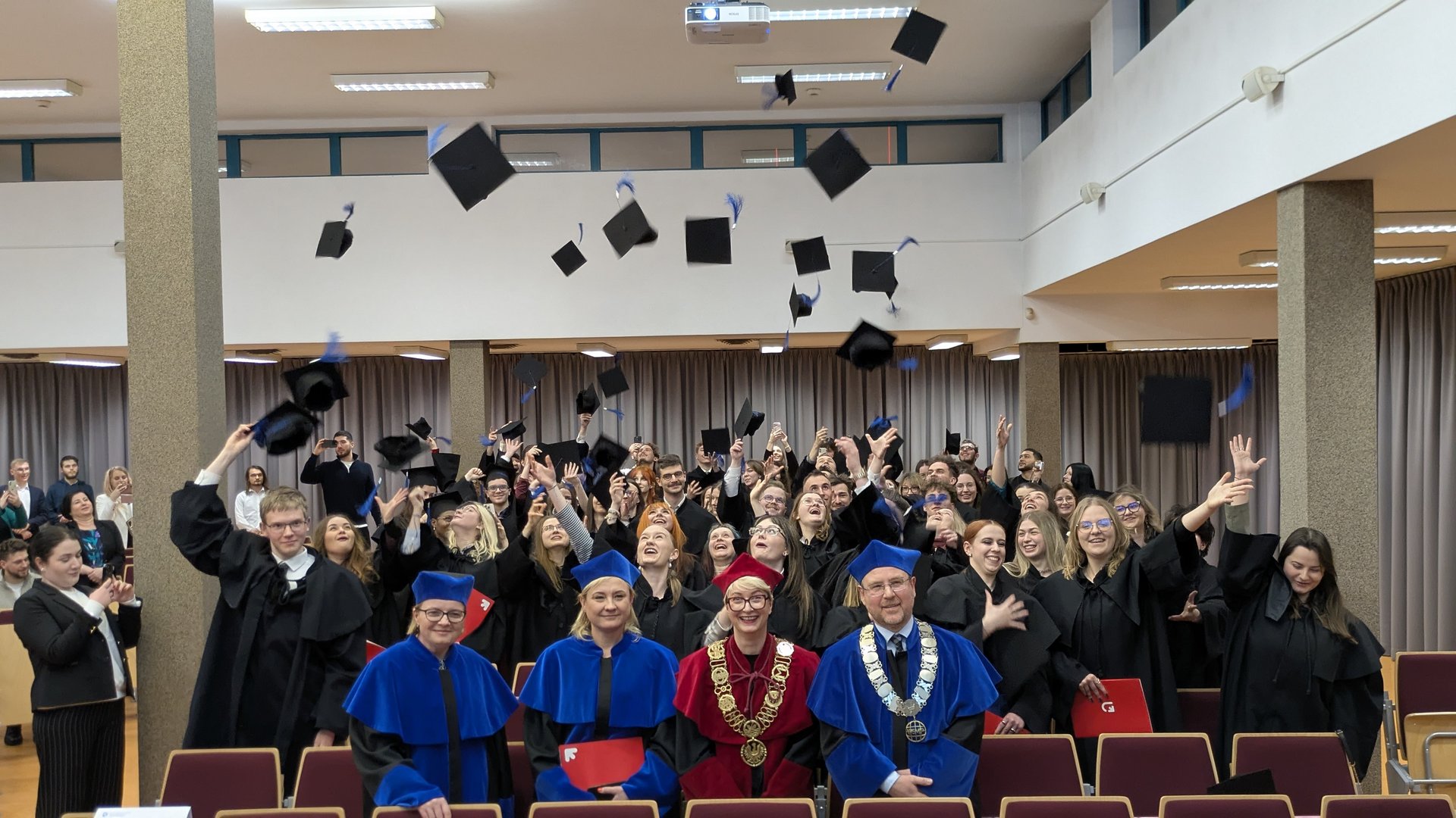 A large group of people in black academic gowns celebrate in a lecture hall by throwing graduation caps into the air; in the foreground stand four individuals in blue and red ceremonial robes, including one wearing a chain of office, with rows of seats, columns, curtains, and ceiling lights visible in the background.