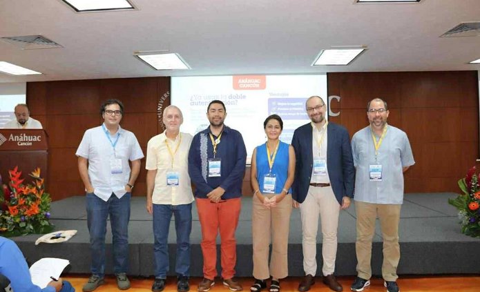 Siedmioro uczestników wydarzenia stoi na scenie w sali konferencyjnej, pozując do wspólnego zdjęcia. Wszyscy mają przypięte identyfikatory, co sugeruje, że są prelegentami lub uczestnikami seminarium. Za nimi widoczny jest duży ekran z prezentacją zatytułowaną ‘¿Es usted el doble cuántico de alguien?’ (Czy jesteś czyimś kwantowym sobowtórem?), z nazwiskami Verónica Santiago i Luis Ignacio Sánchez oraz logo uczelni Anáhuac Cancún. Scena udekorowana jest kwiatami, a po lewej stronie znajduje się mównica z nazwą uczelni.