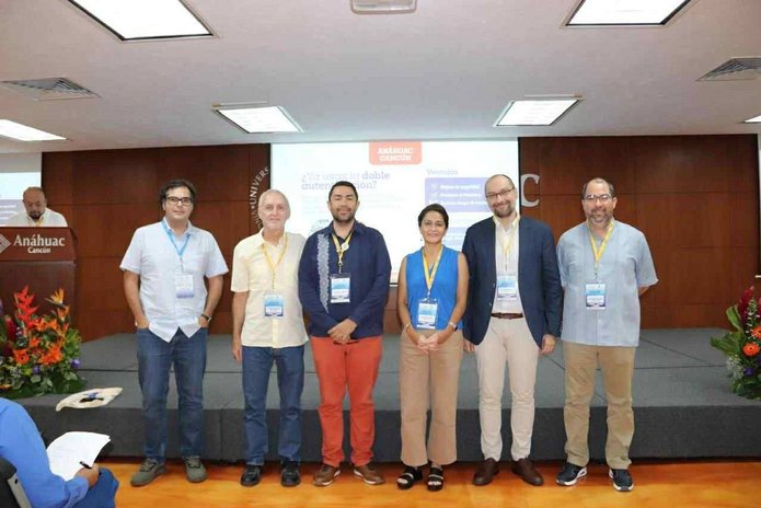 Siedmioro uczestników wydarzenia stoi na scenie w sali konferencyjnej, pozując do wspólnego zdjęcia. Wszyscy mają przypięte identyfikatory, co sugeruje, że są prelegentami lub uczestnikami seminarium. Za nimi widoczny jest duży ekran z prezentacją zatytułowaną ‘¿Es usted el doble cuántico de alguien?’ (Czy jesteś czyimś kwantowym sobowtórem?), z nazwiskami Verónica Santiago i Luis Ignacio Sánchez oraz logo uczelni Anáhuac Cancún. Scena udekorowana jest kwiatami, a po lewej stronie znajduje się mównica z nazwą uczelni.