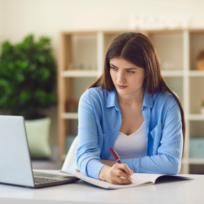 Zdjęcie poglądowe. Dziewczyna spogląda w ekran komputera i robi notatki.