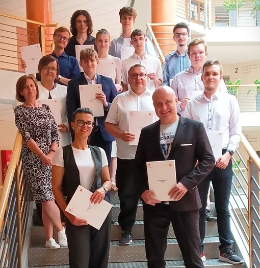 Na zdjęciu znajduje się grupa osób stojących na schodach w budynku. Każda z nich trzyma certyfikat. Osoby są w różnym wieku, elegancko ubrane. W tle widać wnętrze budynku z balustradami i słupami. 