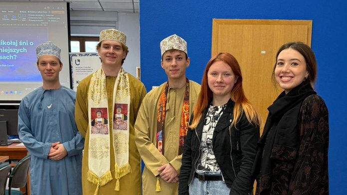 Pięć osób stoi w pomieszczeniu przed niebieską ścianą i drewnianymi drzwiami. Trzy osoby mają na sobie tradycyjne stroje bliskowschodnie, w tym długie szaty i wzorzyste nakrycia głowy. Dwie pozostałe są ubrane w nowoczesne, codzienne ubrania. W tle widoczny jest ekran z prezentacją w języku polskim.