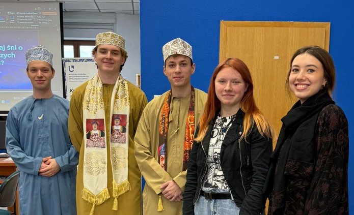 Pięć osób stoi w pomieszczeniu przed niebieską ścianą i drewnianymi drzwiami. Trzy osoby mają na sobie tradycyjne stroje bliskowschodnie, w tym długie szaty i wzorzyste nakrycia głowy. Dwie pozostałe są ubrane w nowoczesne, codzienne ubrania. W tle widoczny jest ekran z prezentacją w języku polskim.