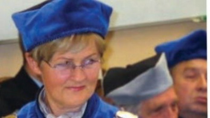 prof. dr hab. Elżbiety H. Oleksy w niebieskiej todze i birecie, z łańcuchem rektorskim, trzyma zielone książeczki lub dyplomy; w tle stoją inni uczestnicy w granatowych togach.