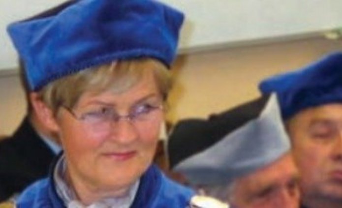 prof. dr hab. Elżbiety H. Oleksy w niebieskiej todze i birecie, z łańcuchem rektorskim, trzyma zielone książeczki lub dyplomy; w tle stoją inni uczestnicy w granatowych togach.