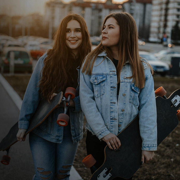Dwie młode kobiety stoją na tle miejskiego krajobrazu z deskorolkami typu longboard w dłoniach. Obie ubrane są w jasnoniebieskie jeansowe kurtki. Jedna z nich uśmiecha się szeroko, a druga patrzy w jej stronę z lekkim uśmiechem. W tle widać zaparkowane samochody i bloki mieszkalne oświetlone ciepłym światłem zachodzącego słońca. Scena emanuje swobodą i miejskim stylem życia.