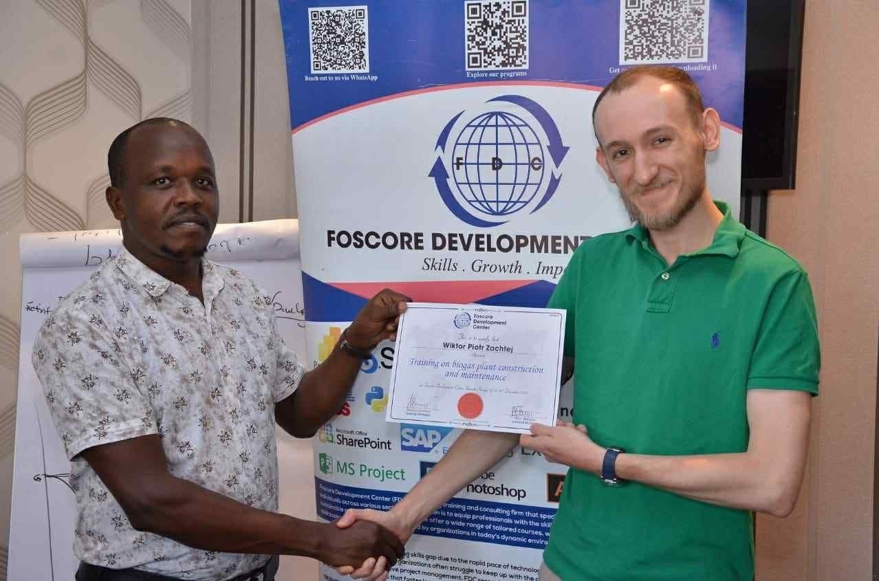 Dwóch mężczyzn podaje sobie ręce podczas wręczenia certyfikatu ukończenia szkolenia z budowy i obsługi biogazowni. Jeden z nich trzyma certyfikat z imieniem Wiktora Piotra Zachleja. Za nimi stoi baner Foscore Development Center z hasłem ‘Skills. Growth. Impact.’ oraz logotypami różnych programów i narzędzi