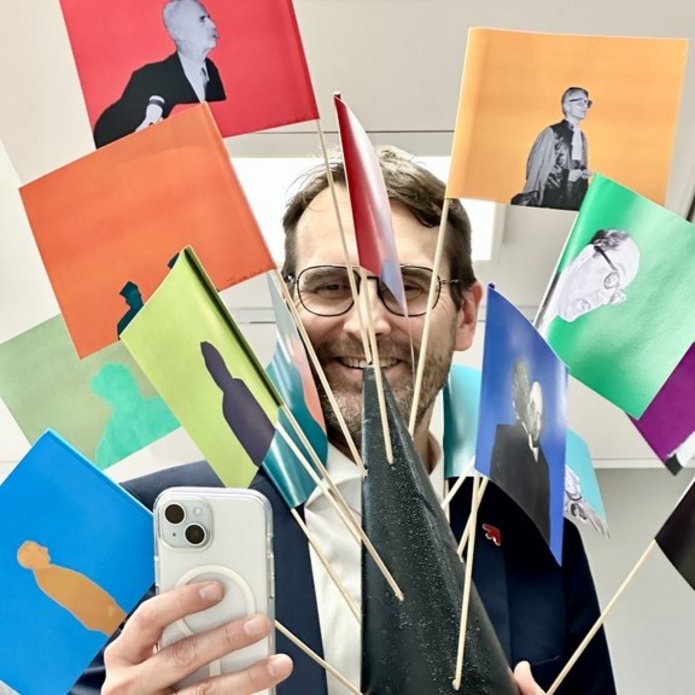 Na zdjęciu widać mężczyznę, który trzyma chorągiewki zatknięte w stożku ze styropianu. Mężczyzna trzyma w drugiej dłoni smartfon i robi nim zdjęcie.