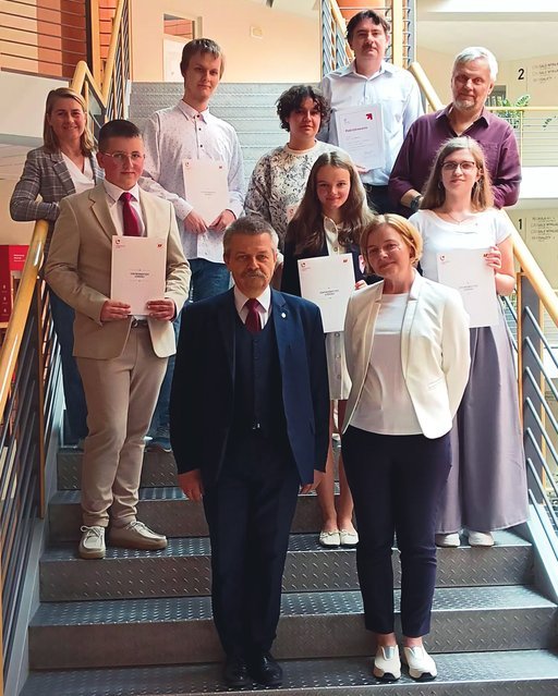 Na zdjęciu znajduje się grupa osób ustawionych na schodach wewnątrz budynku. Wszyscy trzymają certyfikaty i uśmiechają się do kamery. Na pierwszym planie stoi mężczyzna w garniturze i krawacie oraz kobieta w białej bluzce i ciemnych spodniach. Za nimi, na schodach, znajdują się pozostali uczestnicy, ubrani w eleganckie stroje. W tle widać wnętrze budynku z balustradami i słupami.