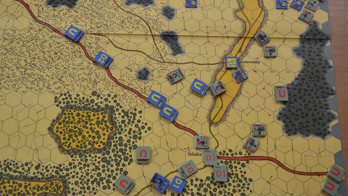 Mapa strategicznej gry planszowej z siatką heksagonalną, przedstawiająca teren z lasami, drogami i wzniesieniami. Na mapie widoczne są oznaczone miejscowości, m.in. „Starawioska” i „Tabalice”. Rozmieszczone są kolorowe żetony jednostek z literami i symbolami, reprezentujące różne frakcje wojskowe — m.in. niebieskie i żółte z oznaczeniami „C” i „P” oraz szare i czerwone z „U” i „D1”. Układ sugeruje rozgrywkę symulującą działania wojenne.