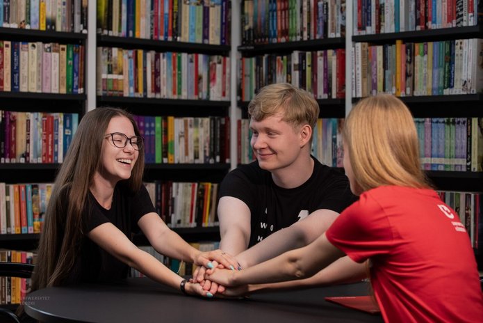 2 studentki i student siedzą w bibliotece przy regałach z książkami i trzymają się za ręce 