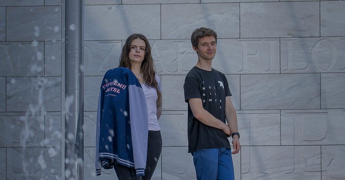 Studentka i student na tle szarej ściany z widocznymi wyrytymi literami. Na pierwszym planie tryskająca woda z fontanny.