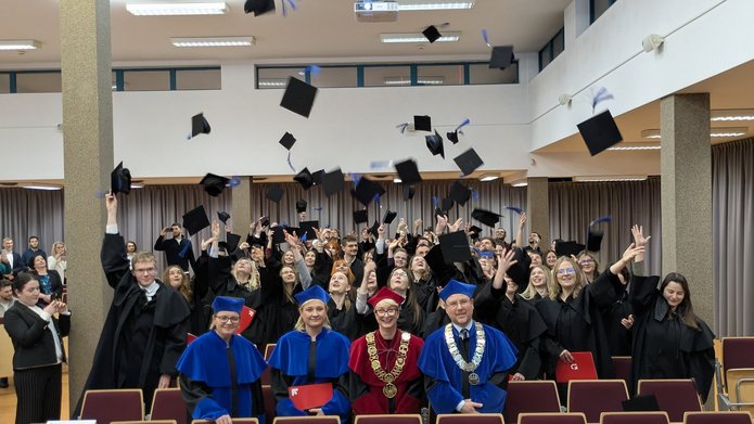 Grupa absolwentów w czarnych togach świętuje w sali, rzucając birety w górę. Na pierwszym planie siedzą trzej akademicy w uroczystych szatach – dwóch w niebieskich i jeden w czerwonej z dużym złotym łańcuchem. W tle widać uśmiechniętych ludzi robiących zdjęcia