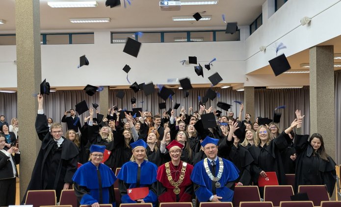 Grupa absolwentów w czarnych togach świętuje w sali, rzucając birety w górę. Na pierwszym planie siedzą trzej akademicy w uroczystych szatach – dwóch w niebieskich i jeden w czerwonej z dużym złotym łańcuchem. W tle widać uśmiechniętych ludzi robiących zdjęcia