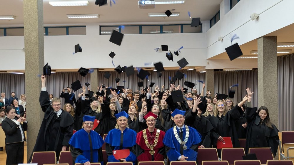 Grupa absolwentów w czarnych togach świętuje w sali, rzucając birety w górę. Na pierwszym planie siedzą trzej akademicy w uroczystych szatach – dwóch w niebieskich i jeden w czerwonej z dużym złotym łańcuchem. W tle widać uśmiechniętych ludzi robiących zdjęcia