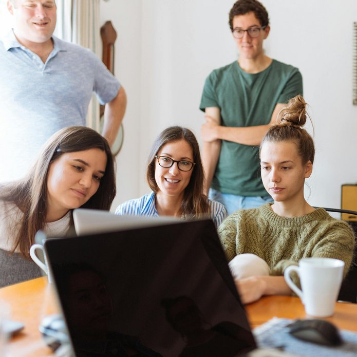 grupa uśmiechniętych młodych ludzi siedzi w pokoju, patrzą na ekran laptopa 