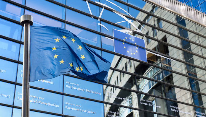 Na pierwszym planie powiewa flaga Unii Europejskiej na maszcie. W tle widoczna jest nowoczesna, przeszklona fasada budynku Parlamentu Europejskiego z napisem powitalnym w różnych językach oraz emblematem UE.