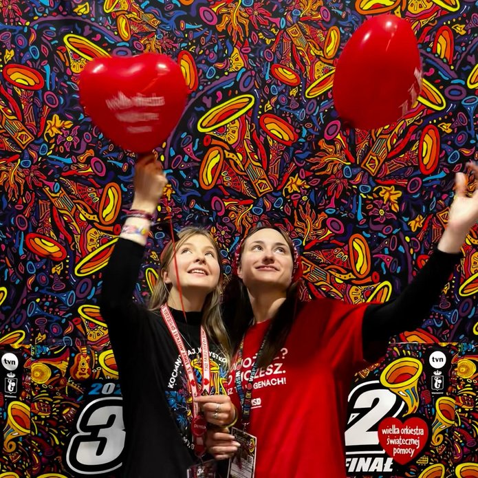 2 young women standing against a colorful background with red heart-shaped balloons with the words The Great Orchestra of Christmas Charity