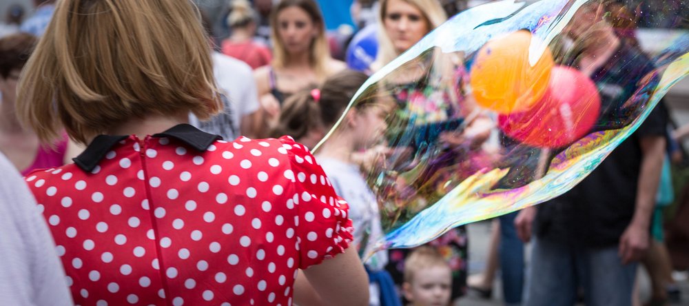 Kobieta w czerwonej sukience w białe groszki z bańkami mydlanymi, w tle widoczne inne osoby